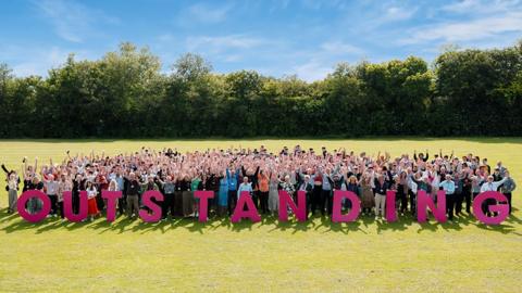 Staff and students celebrating on the college field with sign saying Outstanding