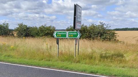 Green road sign at the side of the A15 directing people either towards Sleaford or Lincoln. There is a section of road in view, along with a hedge and a field in the background.