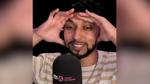 A man with short, dark hair and a short, dark beard holds his hands on his head as he speaks into a microphone.