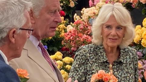 Queen Camilla holds a bouquet of locally-bred peach roses while the King listens to a show official. Behind them is a wall of roses of differing colours and coral alstroemeria flowers. 