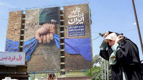 A woman adjusts her headdress as she walks near a billboard on the facade of a building depicting the Strait of Hormuz with a caption in Persian reading "Forever in Iran's Hand", at Vanak Square in Tehran on May 6, 2026. The United States and Pakistan, a key mediator in the Middle East war, expressed new hope of talks leading to a peace deal with Iran on May 6, after President Donald Trump halted a plan to guide ships through the Strait of Hormuz. 