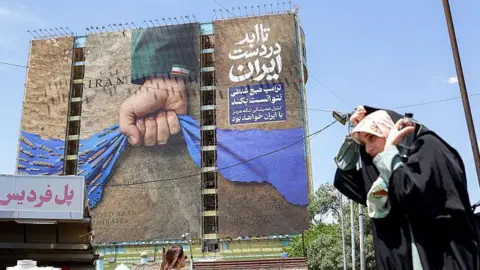A woman adjusts her headdress as she walks near a billboard on the facade of a building depicting the Strait of Hormuz with a caption in Persian reading "Forever in Iran's Hand", at Vanak Square in Tehran on May 6, 2026. The United States and Pakistan, a key mediator in the Middle East war, expressed new hope of talks leading to a peace deal with Iran on May 6, after President Donald Trump halted a plan to guide ships through the Strait of Hormuz. 