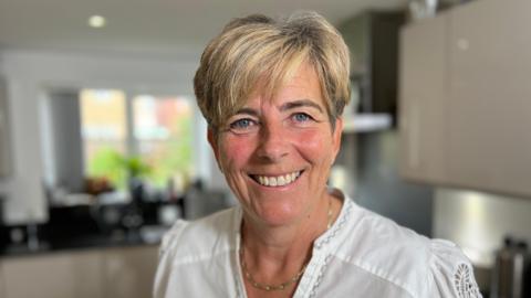 Cancer survivor Lisa Noble with short blonde highlighted hair and blue eyes. She is wearing a white top and gold necklace. She is smiling into the camera while standing in a kitchen, which is blurred in the background.