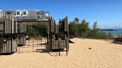 A park on sand. There is a wooden climbing frame. Behind the park is a house. The sky is blue. In the background to the right is the sea and sailing boats. 