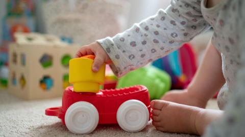 A toddler plays with a red plastic toy car while sitting in a bedroom.