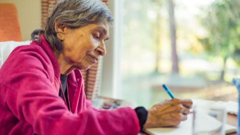 A stock image of an elderly woman writing on a note book. She is wearing a dark pink cardigan and black t-shirt and is using a blue pen. 