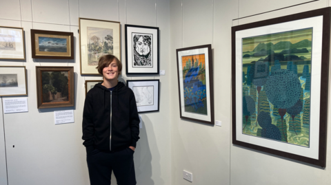 Valya Constable smiles for the camera next to his framed artwork in the exhibition