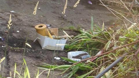 A ripped cardboard box containing grenades and mines sits on dry, muddy ground edges by unkempt green plants