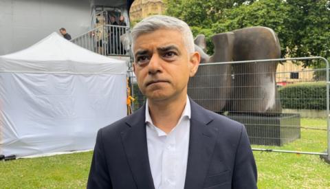 The London Mayor is wearing a suit and shirt, looking slightly off camera. He is stood in front of a sculpture to his left and white tent to his right. 