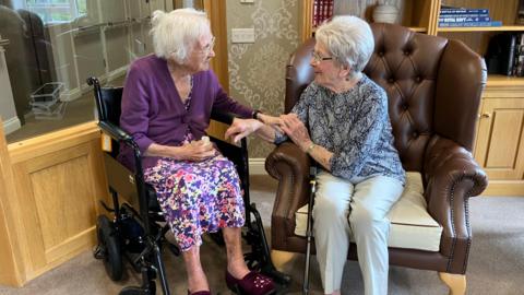 Two women speak to each other - both have white hair and one is wearing a grey pattern shirt and grey trousers and sits on a brown armchair while the other is in a wheelchair and wears a floral skirt and purple cardigan