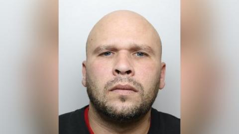 A mugshot of a bald man who has a beard.