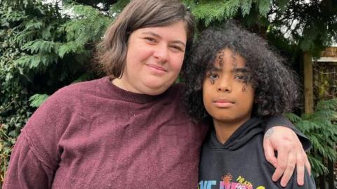 Glyn and Kymani are stood side by side in their garden and smiling slightly. Glyn is wearing a dark red woollen jumper, and has chin-length dark brown hair - she has her arm around her son's shoulders. Kymani is wearing a black hoodie with 'One Piece' written on it in yellow, blue and pink writing. He has shoulder-length, curly black hair. Behind the pair is a dark green evergreen tree, with a wooden garden fence to the right of the photograph. 