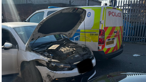 A white car stands next to a police van. The white car's bonnet is open and there are scuffs along the front of the car plus damage along the side.