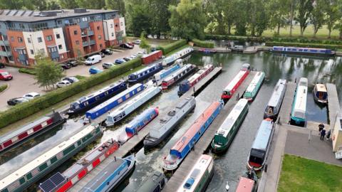 Drone shot shows a marina with rows of narrowboats docked. A residential building is nearby with parked cars on the left.


