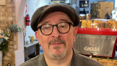 Daren Wayland stands in his coffee/barber shop and faces the camera. He wears a dark green flat cap, with black round glasses and a green shirt. He has a piercing in his left ear. The coffee shop can be seen behind him.