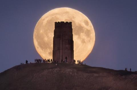 A group of people stand on Glastonbury Tor at night with a giant supermoon framing the monument on the top behind them