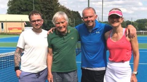 Three men and a woman stand together on a tennis court and smile at the camera. They are wearing sports clothes.
