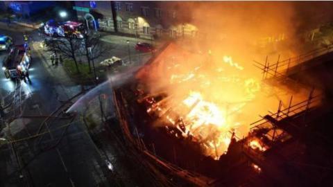 An aerial view of the Wesley chapel on fire. Flames are engulfing the roof of the structure. A large stream of water is coming from a fire engine to extinguish the blaze.