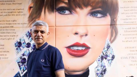 Sadiq Khan smiling at the camera, wearing a navy Fred Perry t-shirt, stook in front of a large murial of Taylor Swift with writing around her face. In the artwork, Taylor has red lipstick on and large diamond-shaped earrings with purple gems. 