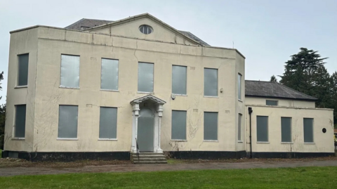 An old cream coloured two-storey building, with lots of windows, that are all sealed with metal covers. There is a side wing, and grass at the front. The front door also has a metal cover and has four steps leading up to it. 
