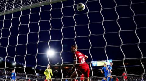 A football match is taking place on a dark night. A ball is being kicked into the net and players are watching it in the air. We can see a floodlight shining bright and fans in the background. There is also a bright screen showing the score and the time. 