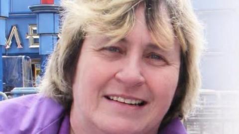 A close up of Theresa Wheelhouse. She has shoulder length blonde/brown hair and is wearing a purple jacket.