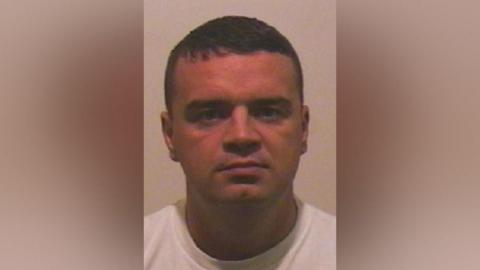 Mugshot of Monaghan. He is clean shaven with short dark hair and wears a white t-shirt.