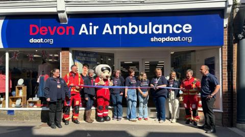 A ribbon-cutting ceremony in front of a store a newly opened Devon Air Ambulance charity shop. There are 11 people stood outside the shop all wearing company uniform. One is dressed as a mascot. 