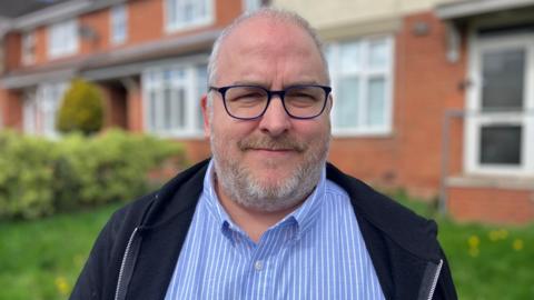 A man with short grey hair and a grey beard, wearing blue-rimmed spectacles, a blue-and-white-striped shirt and a dark blue jacket, stands in front of some out-of-focus houses.