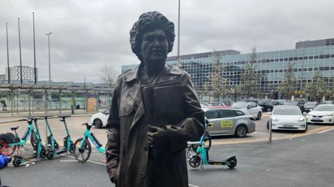 A life‑sized statue of Jennie Lee MP wearing a long coat and holding a folder against her chest. The statue stands outdoors in an urban setting near a row of bicycles and scooters, with parked cars and modern buildings visible in the background under an overcast sky.