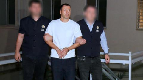 A man in a white T-shirt is held by the arms by two men in dark-coloured uniform