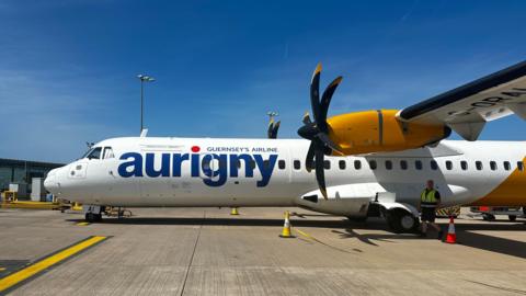 Side view of an Aurigny ATR aircraft on an airport apron under a clear blue sky, with yellow engine and black propellers visible.