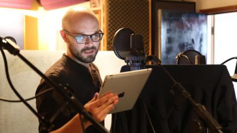 Peter stands talking in front of a microphone. He is reading from a script on his iPad whilst wearing a black t-shirt. He is bald.