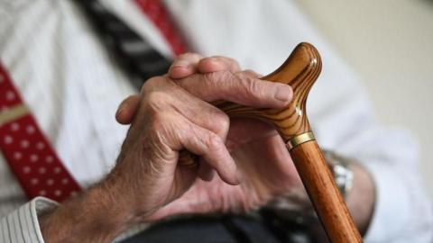 A close of an elderly man's hands. He is holding a walking stick and wearing a white shirt and a tie