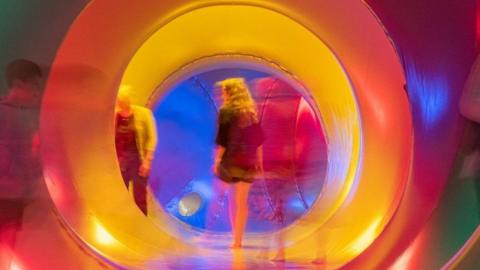 A woman walks through the Luminaire. She is walking through a brightly lit tunnel with a mix of colours including red, orange, yellow and blue.