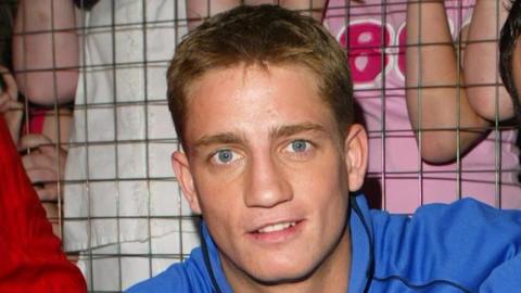 Philip Olivier is pictured during his Brookside years while on SMTV Live. He is wearing a blue hooded jumper with the words "Brookside team". He has short blonde hair and is smiling at the camera. 