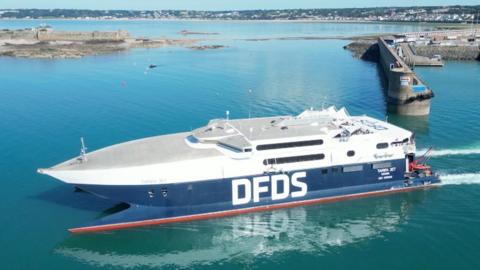 A white and blue ferry with the logo DFDS in white along the side is sailing in St Helier. It is s bright sunny day and the water is turquoise.