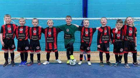 Young girls and boys standing in a row, in a sports hall. They are in front of a football goal and have their arms around each other's shoulders. They are all wearing a red and black striped football kit. 