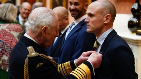 King Charles the third presenting a medal to Dr Matthew Newport. Newport has no hair and is wearing a navy suit.