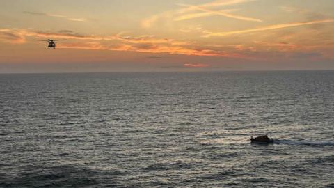 A helicopter and a lifeboat search the sea for a missing person during sunset.