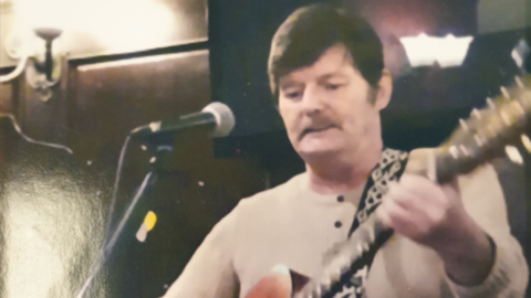 A man with short black hair and a beige jumper stands in front of a microphone while holding a guitar.