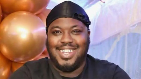 Mr Smith is smiling at the camera. He has a black cloth or cap tied tightly to his head and he has a close cropped beard.