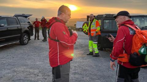The picture shows a group of people outdoors during sunset, standing near several vehicles. Many of the people are dressed in outdoor gear, with some wearing red jackets and one person in high-visibility clothing. 