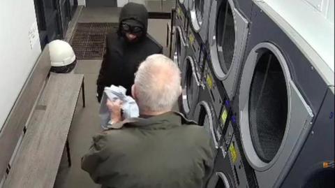 A CCTV image showing a man wearing a green jacket holding a pair of blue denim jeans, facing a man wearing a black coat and black mask. On the right are double-stacked washing machines and driers