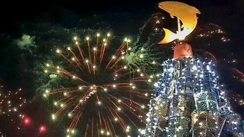 A brightly lit Christmas tree with a large gold salmon cut out on top and a sky lit with fireworks.