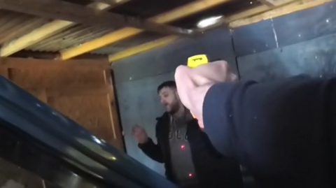 Reece Reddington in a garage with his hands up, and a taser being pointed at him