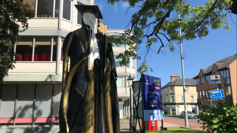 A picture of the Cambridge Don statue. It depicts a white statue wearing a university robe and hat in black and gold. It sits in front of a building near trees. Other buildings and a road can be seen in the background.