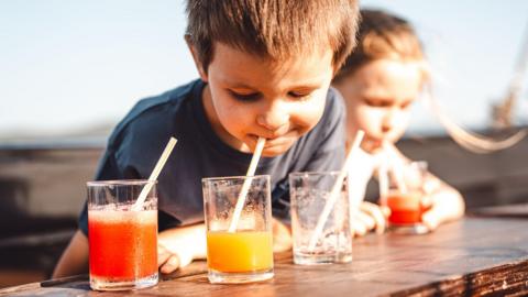 A young boy drinks from a glass of juice though a straw while sitting outdoors at a picnic bench in the sun with a friend