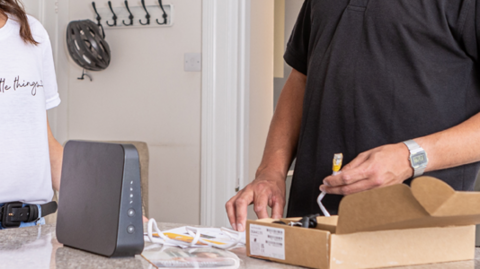 A Sure Guernsey engineer wearing a black polo shirt and baseball cap with the company's logo on it connects a black broadband router in a woman's kitchen while she watches on and chats to him. The woman is wearing a white t-shirt and blue jeans and has long brown hair. A green clock on wall says the time is 12:15 and a cycling helmet is on a clothes hook.