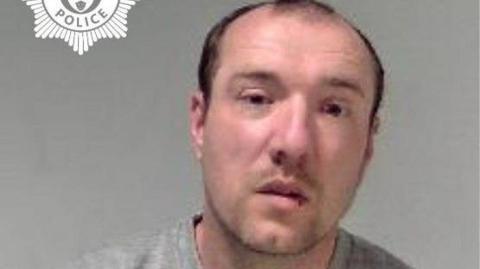 A man with black receding hair is photographed in a grey sweatshirt in front of a grey background.